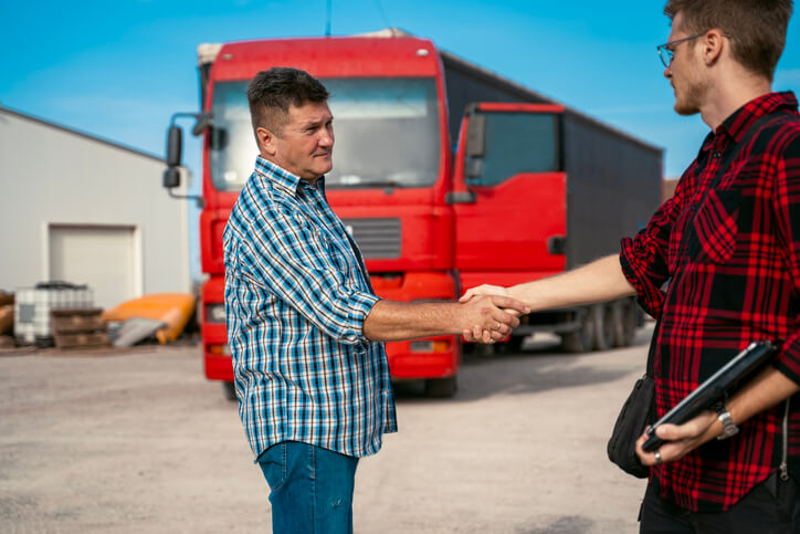 A male driver recruiter holding a notepad while shaking the hands of a truck driver at a warehouse after dispatch college