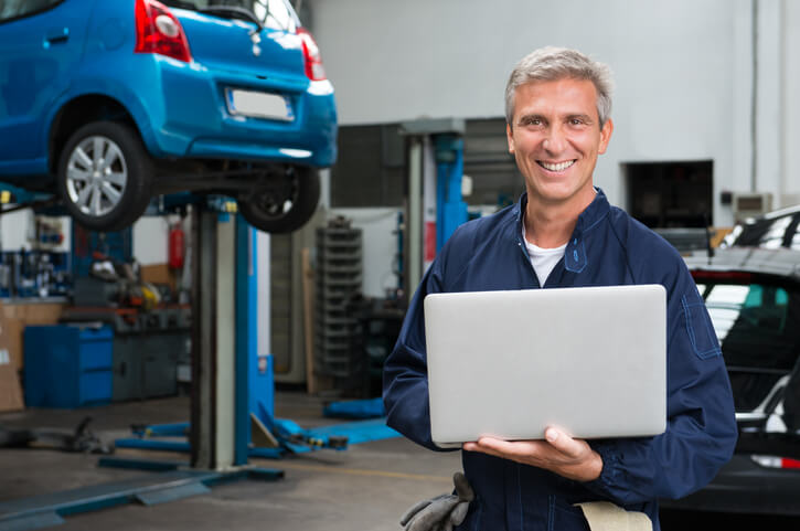 A smiling male auto mechanic holding a laptop in a repair shop after completing auto mechanic training