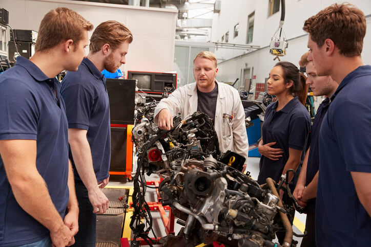 A group of apprentice students interacting with an experienced mechanic at an automotive school