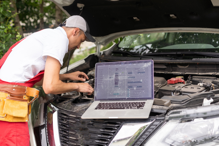 Male electrical specialist working close to an automobile engine while using a laptop after completing his auto mechanic training