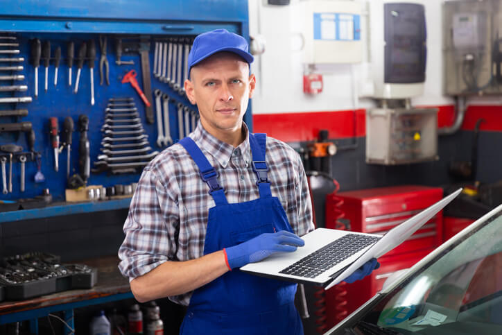 A confident male mechanic holding a laptop at a repair shop after graduating from auto mechanic school