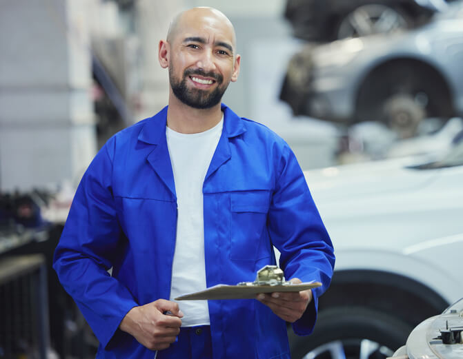 A smiling male auto mechanic holding a clipboard at a repair shop after completing his auto mechanic training