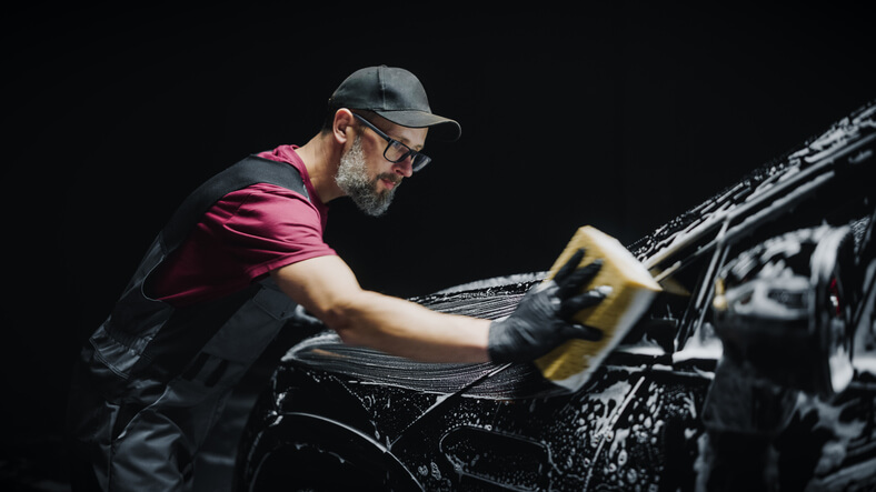 A professional auto detailing specialist using a big soft sponge to wash a high-tech black family electric SUV with foam before detailing, polishing, and waxing