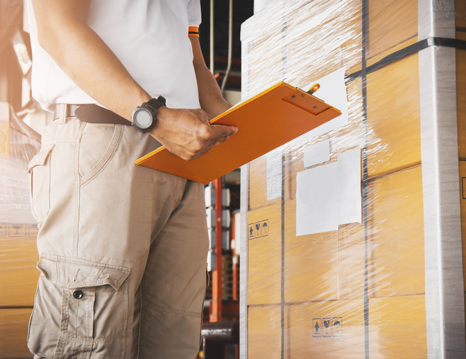 Dispatcher holding a clipboard and managing inventory in a warehouse, emphasizing the importance of detail-oriented skills in auto careers and logistics.